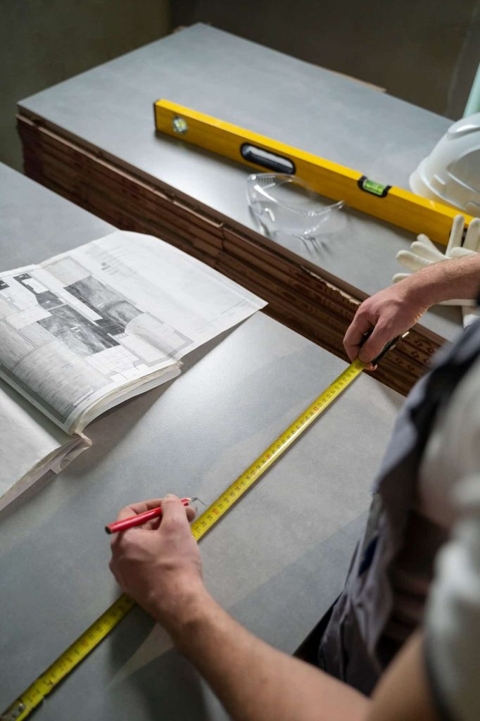 A worker measuring a surface with a tape measure and marking it with a pencil, alongside blueprints, a level tool, and safety gear on the table