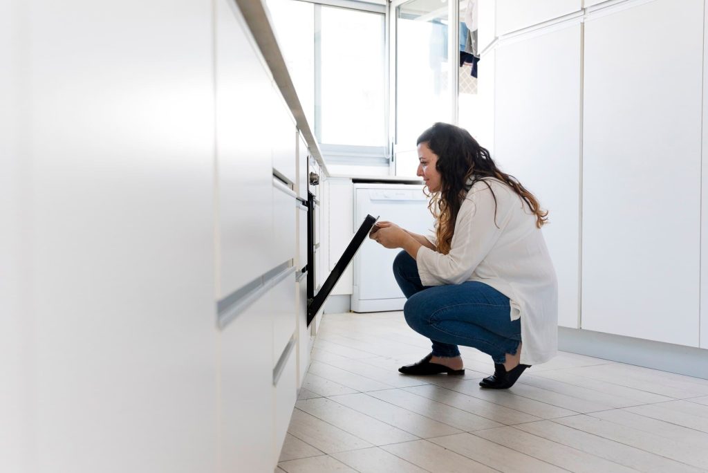 A woman inspecting an open oven in a bright, modern kitchen with white cabinets and tiled flooring