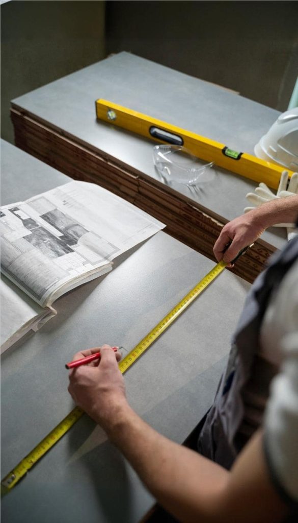 Worker measuring a surface with a tape measure for accurate tile layout planning, alongside As-Built documentation and tools.